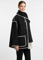 Frau trägt einen schwarzen Poncho-Schalmantel mit weißen Fransen und kontrastierenden Nähten vor weißem Hintergrund