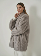 Frau trägt eine graue Strickjacke mit Zopfmuster vor einfarbigem Hintergrund