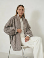 Frau trägt eine graue Plüschjacke über einem weißen Oberteil und einer Hose und sitzt auf einem Stuhl vor einfarbigem Hintergrund