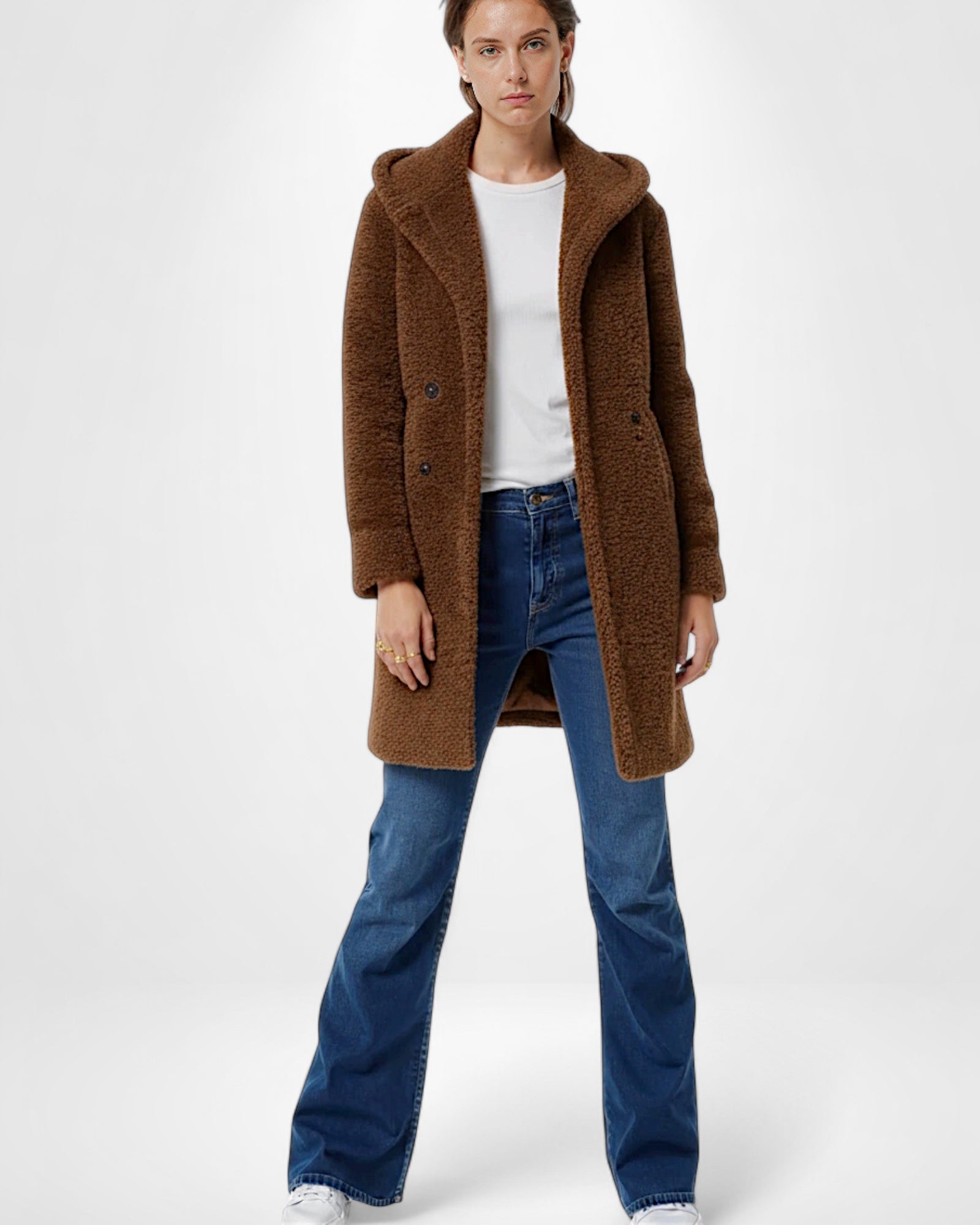 Frau trägt einen braunen Teddyfellmantel, ein weißes Hemd und eine blaue Jeans vor weißem Hintergrund