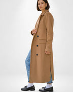 Frau trägt einen mittelangen, hellbraunen Mantel über Jeans vor weißem Hintergrund