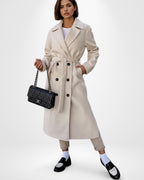 Frau trägt einen beigefarbenen Winter-Trenchcoat mit Gürtel und hält eine schwarze Handtasche vor weißem Hintergrund