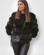 Frau trägt einen schwarzen Webpelzmantel mit passender Tasche vor weißem Hintergrund