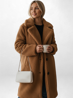 Frau trägt einen braunen Teddy-Mantel und hält eine weiße Tasse sowie eine weiße Handtasche vor weißem Hintergrund