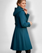 Frau trägt einen knielangen petrolblauen Mantel mit Kapuze vor hellgrauem Hintergrund