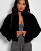 Frau trägt eine schwarze Teddyfelljacke über einem grauen Outfit vor weißem Hintergrund