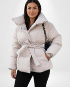 Frau trägt eine beige Pufferjacke mit Gürtel vor weißem Hintergrund
