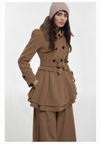 Frau trägt einen khakifarbenen Wintertrenchcoat mit gefüttertem Kragen und einen Hut vor weißem Hintergrund