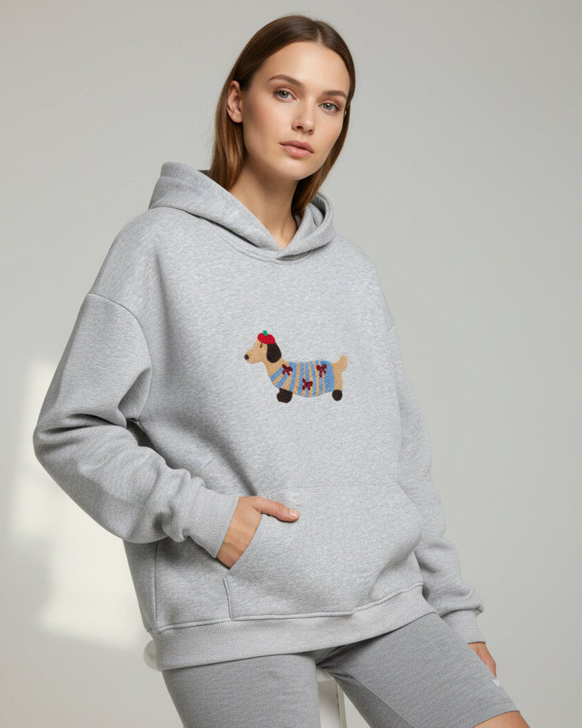 Junge Frau trägt einen hellgrauen Hoodie mit Dackelmotiv vor schlichtem Hintergrund