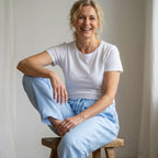 Eine Frau sitzt auf einem Holzhocker und trägt ein weißes T-Shirt sowie eine hellblaue Leinenhose