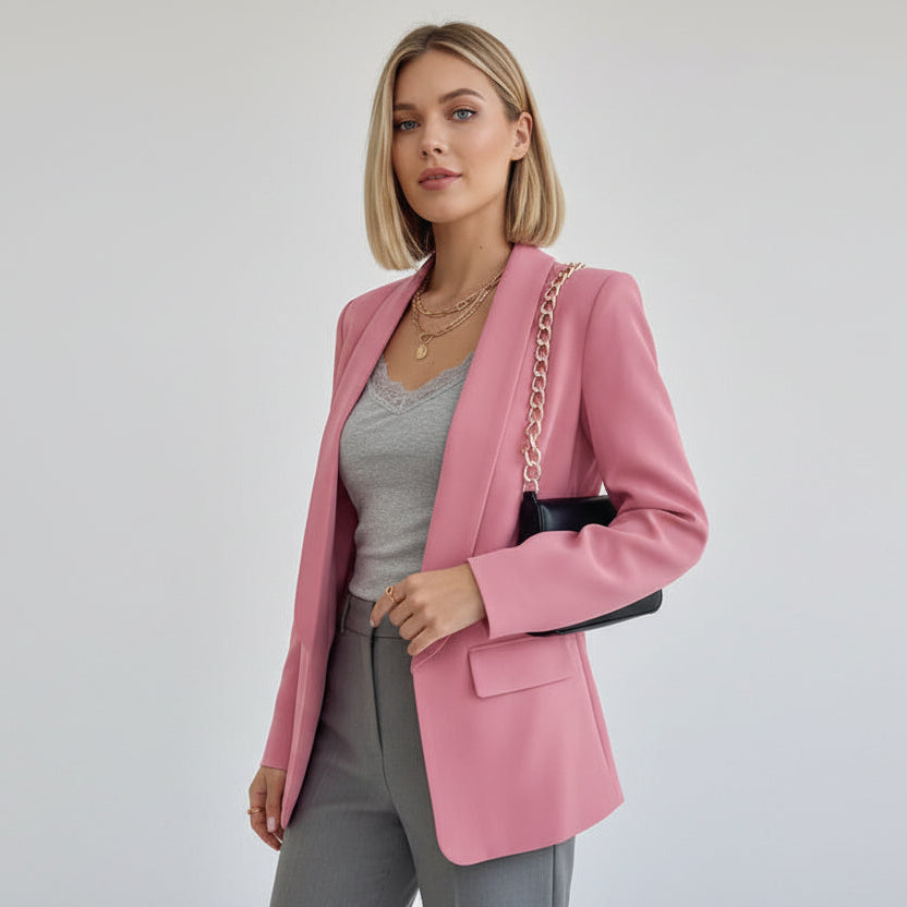 Frau trägt einen rosa Blazer über einem grauen Outfit vor schlichtem Hintergrund.